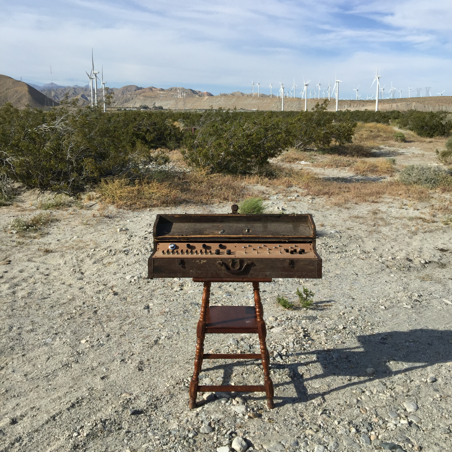 The, a wooden toolbox with a inlayed wooden interface covered in buttons, switches and knobs. The instrument appears to be almost keyboard-esque. It is resting on a wooden stand in the desert outside Palm Springs. In the distance you can see mountains and windmills